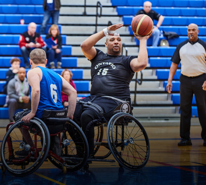 Two basketball players on the basketball court. One player is shooting the ball in the air while the other to the left is trying to block them from shooting.